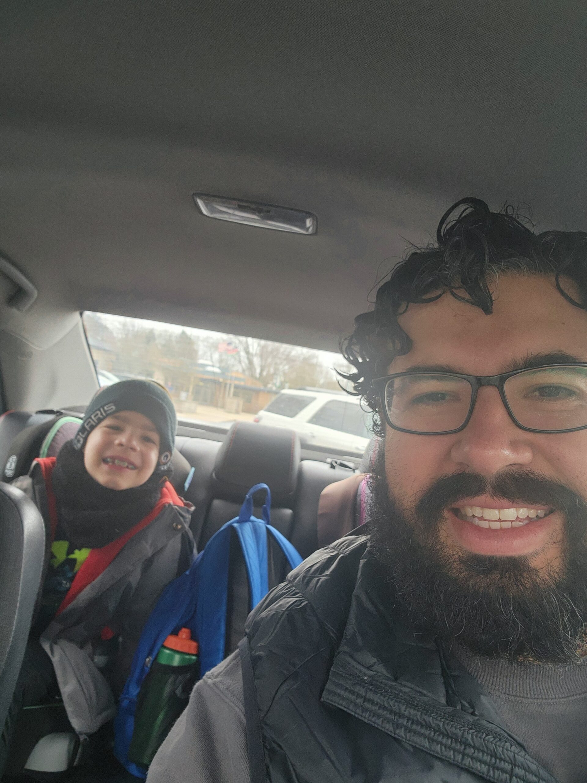 An adult and a child sit in a car. The child is smiling in the back seat with a backpack, and the adult is in the front taking a selfie.