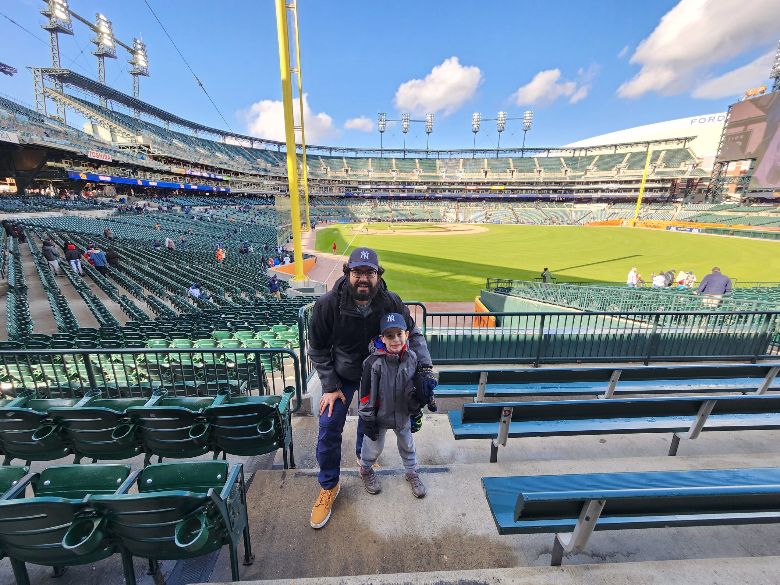A father and son wearing baseball caps pose for a photo in an empty baseball stadium, capturing a special Father-Son Morning lessons moment with the field and stands visible in the background.