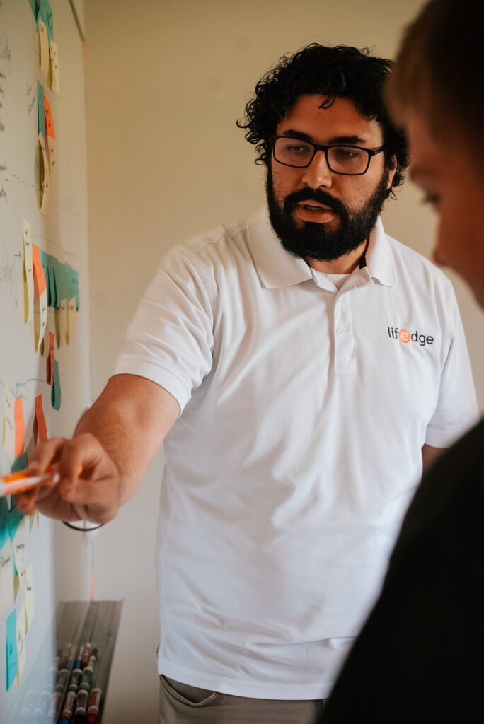 A man in a white "lifedge" polo shirt points at sticky notes on a whiteboard while speaking to another person.