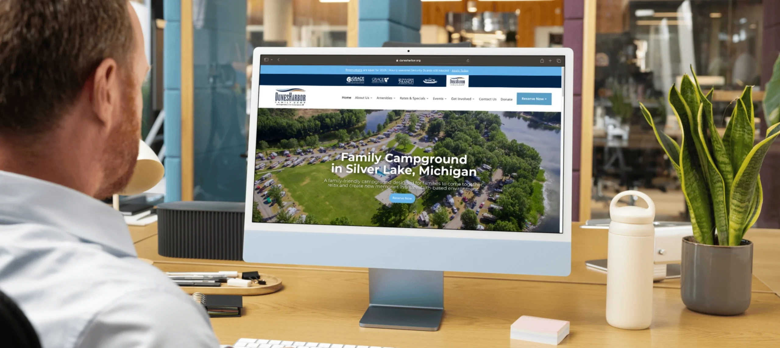A person sits at a desk viewing a campground website on a desktop computer screen in a modern office setting.