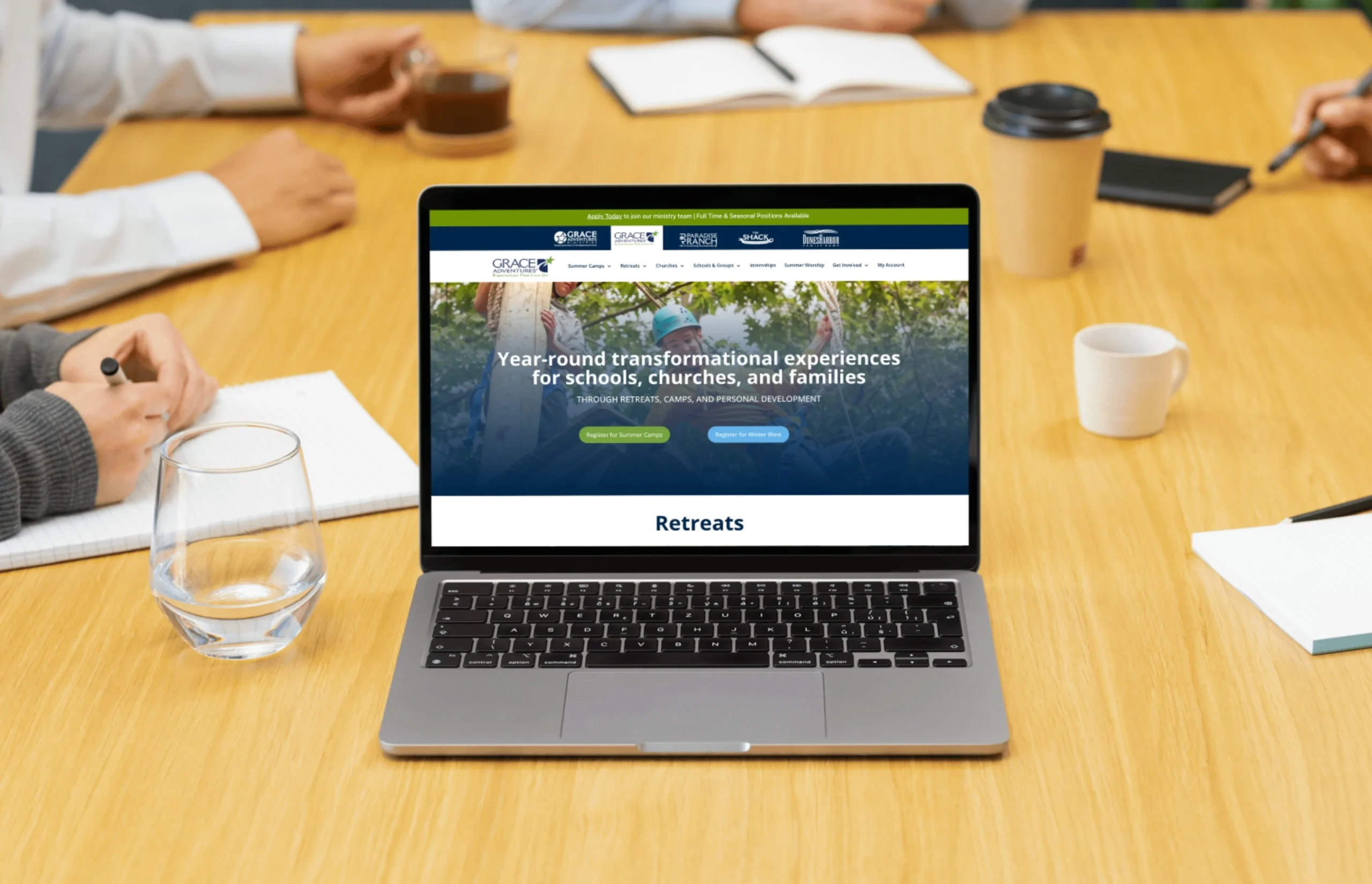 A laptop displaying a website for retreats is on a meeting table with notebooks, pens, coffee cups, and people in the background.