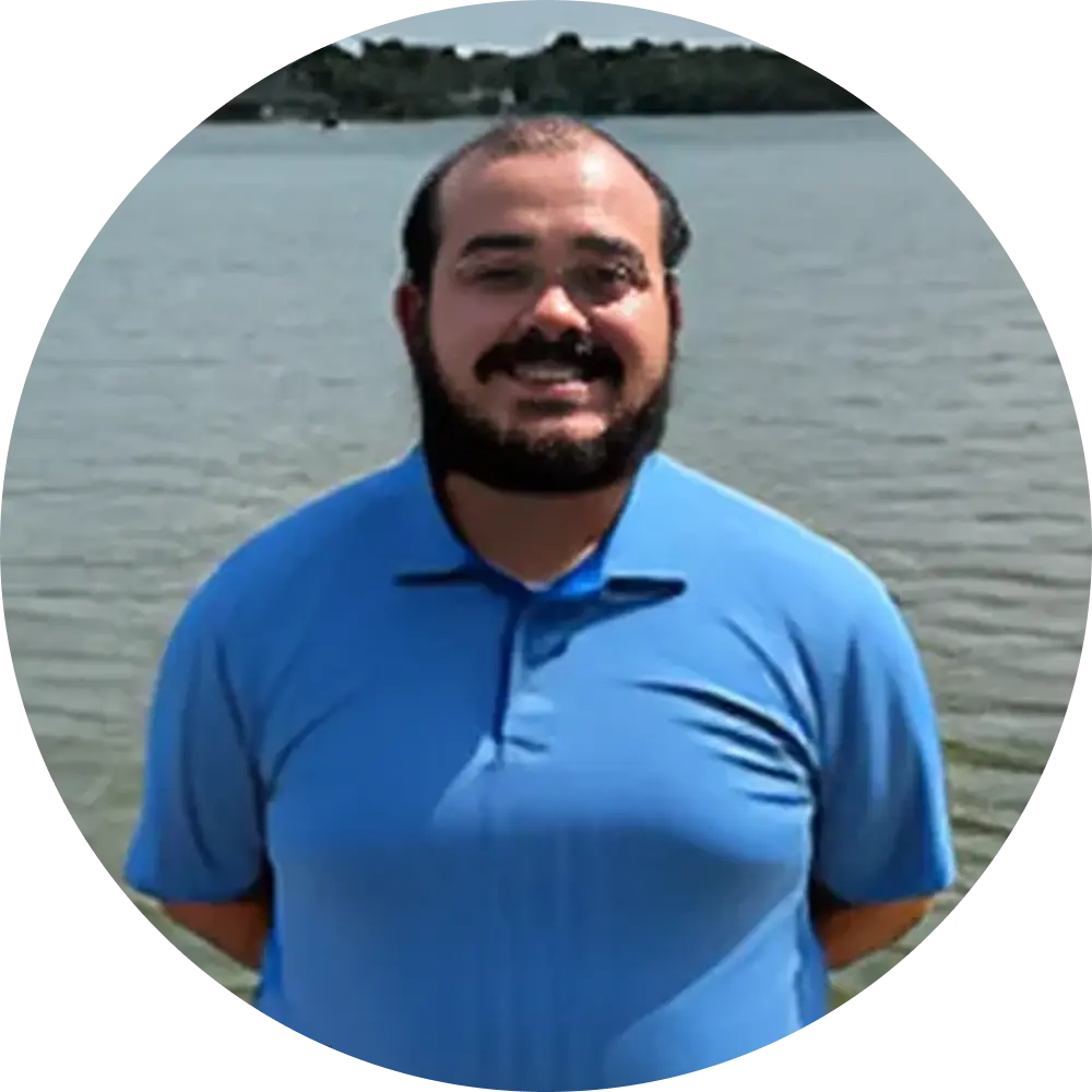 A man with a beard wearing a light blue polo shirt stands in front of a body of water, smiling at the camera.