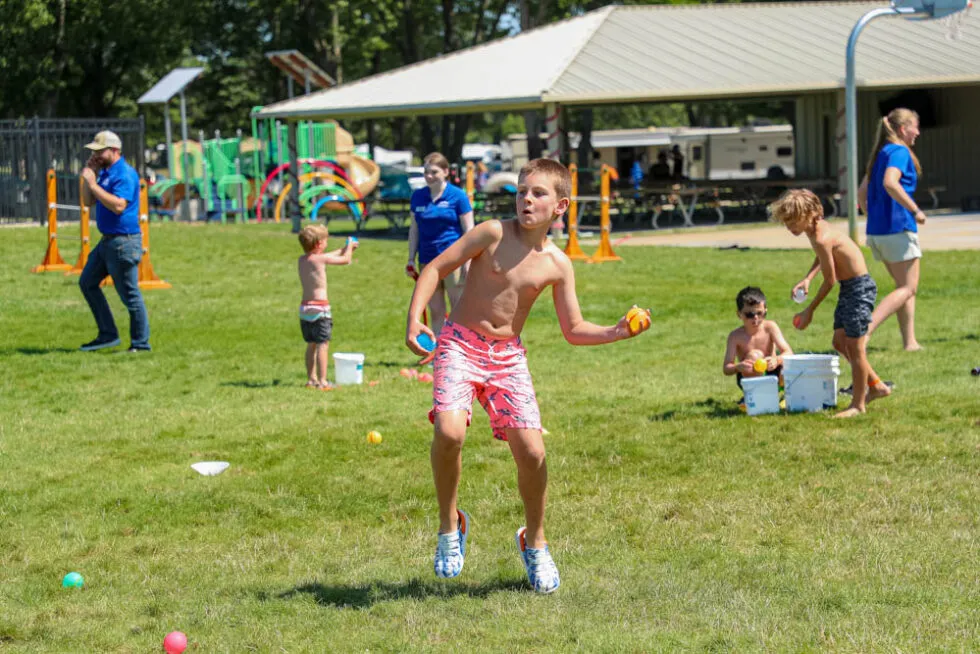 Children and adults play with water balloons and coolers on a grassy field near a playground and pavilion on a sunny day.