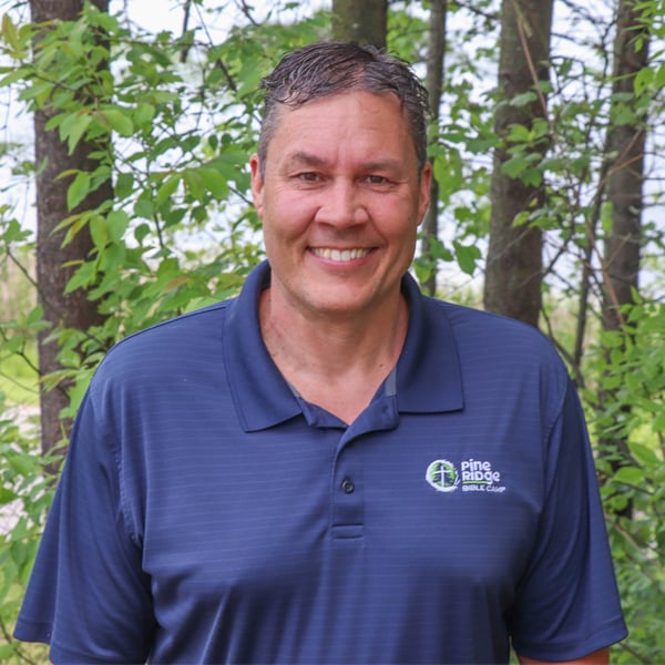 A man wearing a blue Pine Ridge Resort & Club polo shirt stands outdoors in front of green trees, smiling at the camera.