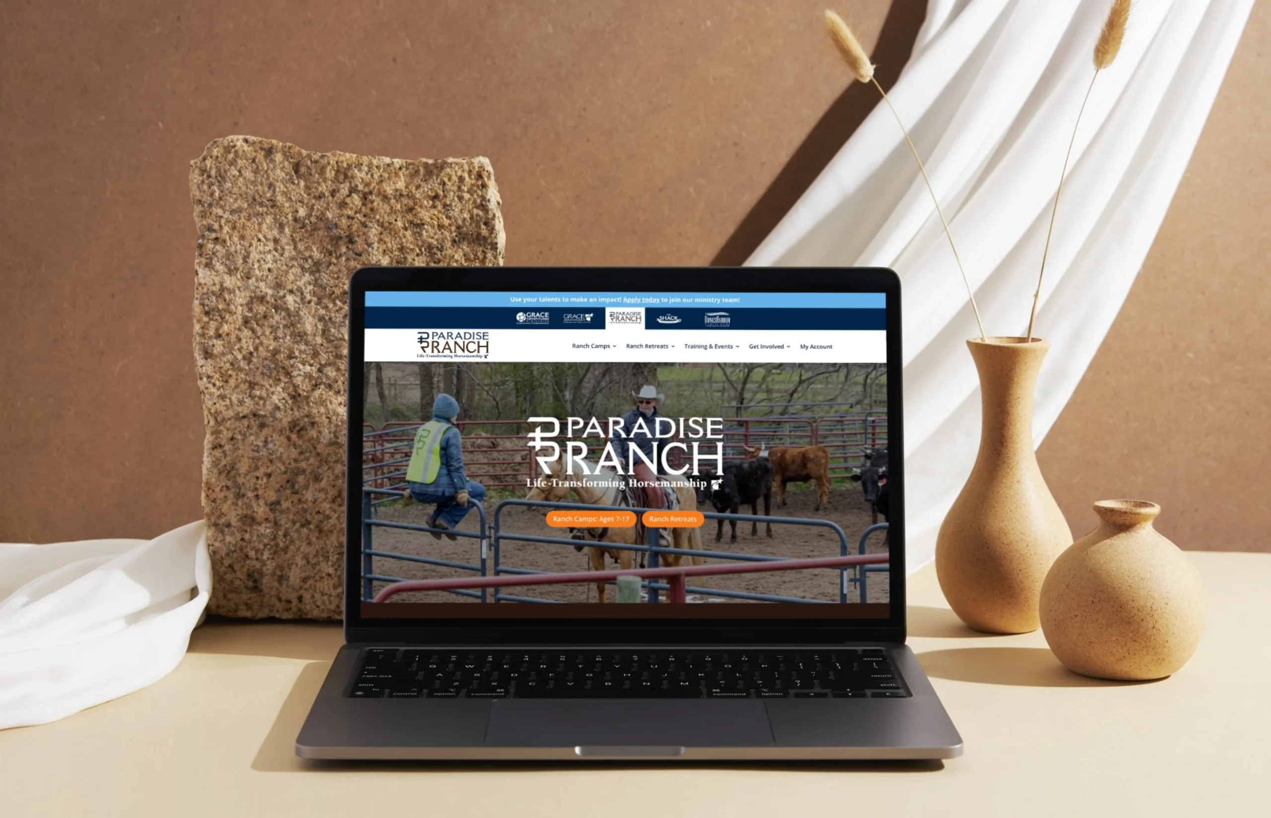 A laptop on a beige surface displays the Paradise Ranch website, with two vases and a stone in the background against a beige draped cloth.