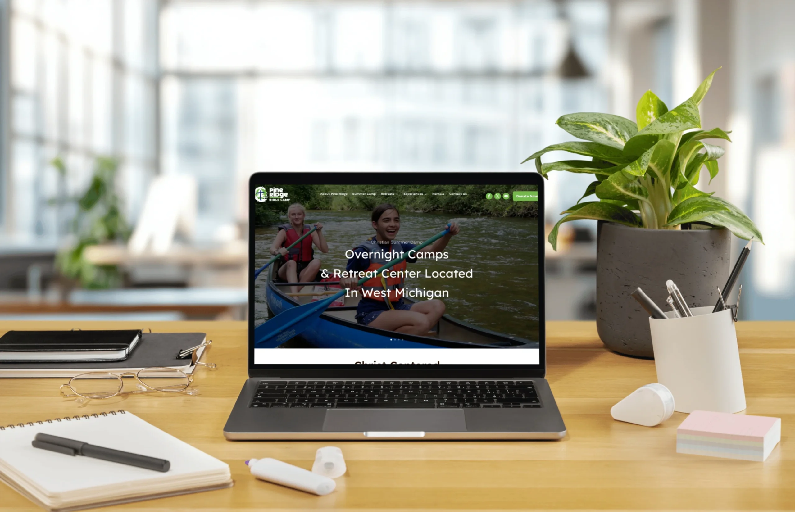 A laptop on a wooden desk displays a website for overnight camps and retreat centers, with a blurred modern office background and various desk items nearby.