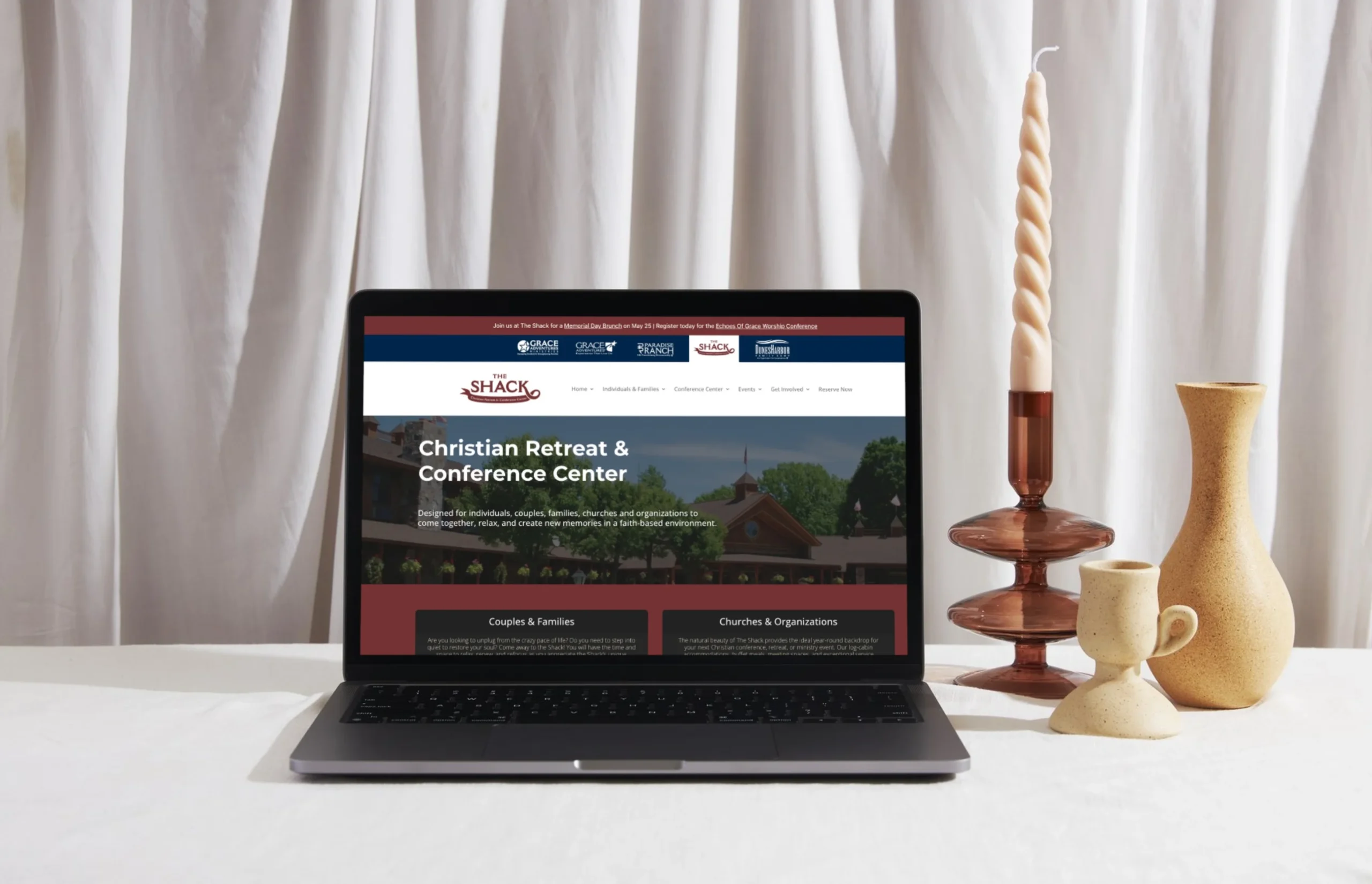 A laptop displaying the website for a Christian Retreat & Conference Center sits on a table with a brown candlestick and twisted candle, against a white curtain backdrop.