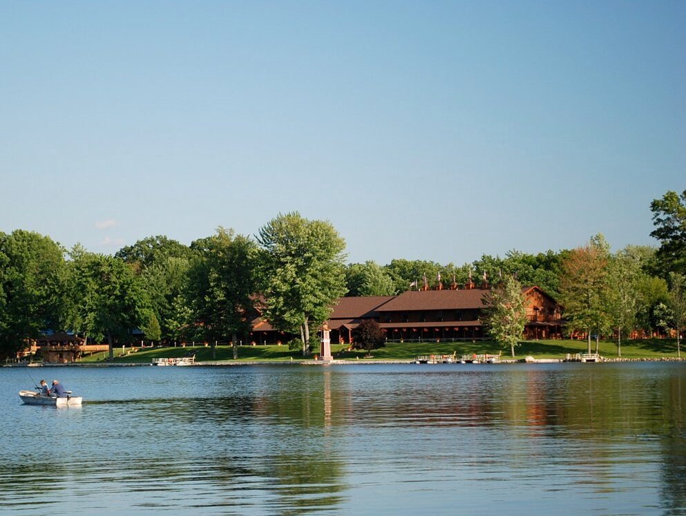 A calm lake with people in a small boat near the shore, trees, and a large wooden building in the background under a clear blue sky.