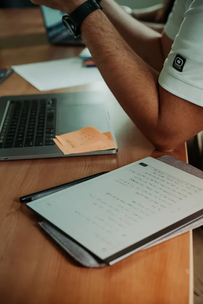 A person sits at a desk with a laptop, sticky notes, and a tablet showing handwritten notes.