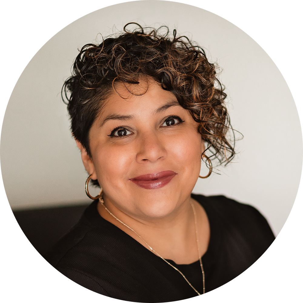 A woman with short, curly hair and hoop earrings smiles at the camera, wearing a black top and a necklace. The background is plain and neutral.